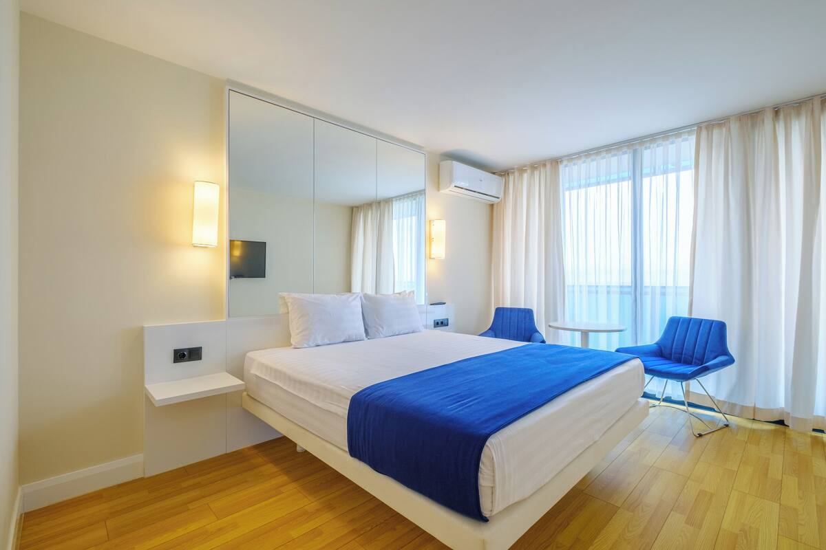 Una habitación de hotel moderna con una cama grande, una silla azul, una mesita pequeña y un espejo amplio.