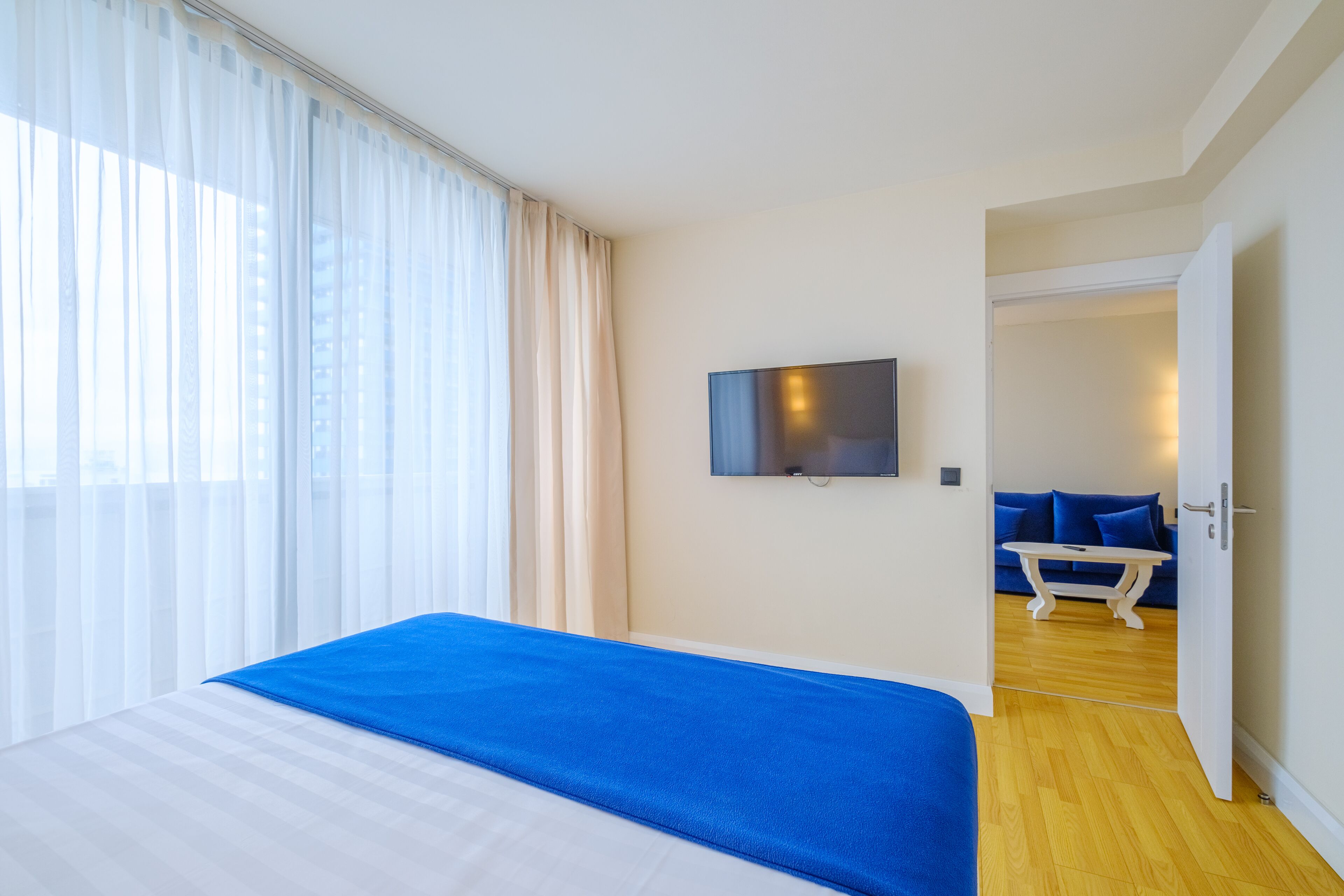 A hotel room with a bed, a TV mounted on the wall, and a view of a blue sofa and table through an open door.