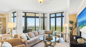 A spacious living room with a large sectional sofa, a glass coffee table, and a view of the ocean through large windows.