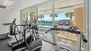 A fitness area with treadmills and an elliptical machine, overlooking a pool with palm trees and a clear sky. - Luxury living at the sea for up to 8 people! | Blue Water Keyes - PH 9 (North Myrtle Beach)
