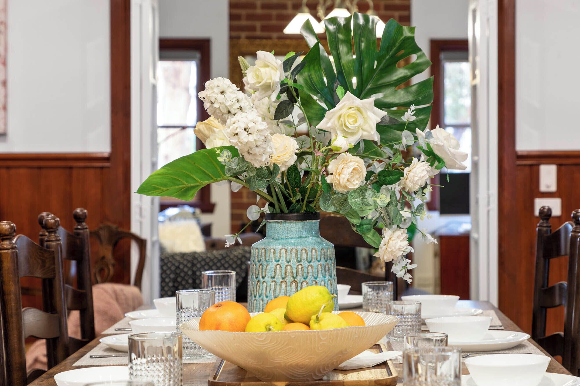 A dining table set with white plates, clear glasses, a vase with white flowers, and a bowl of fruit.
