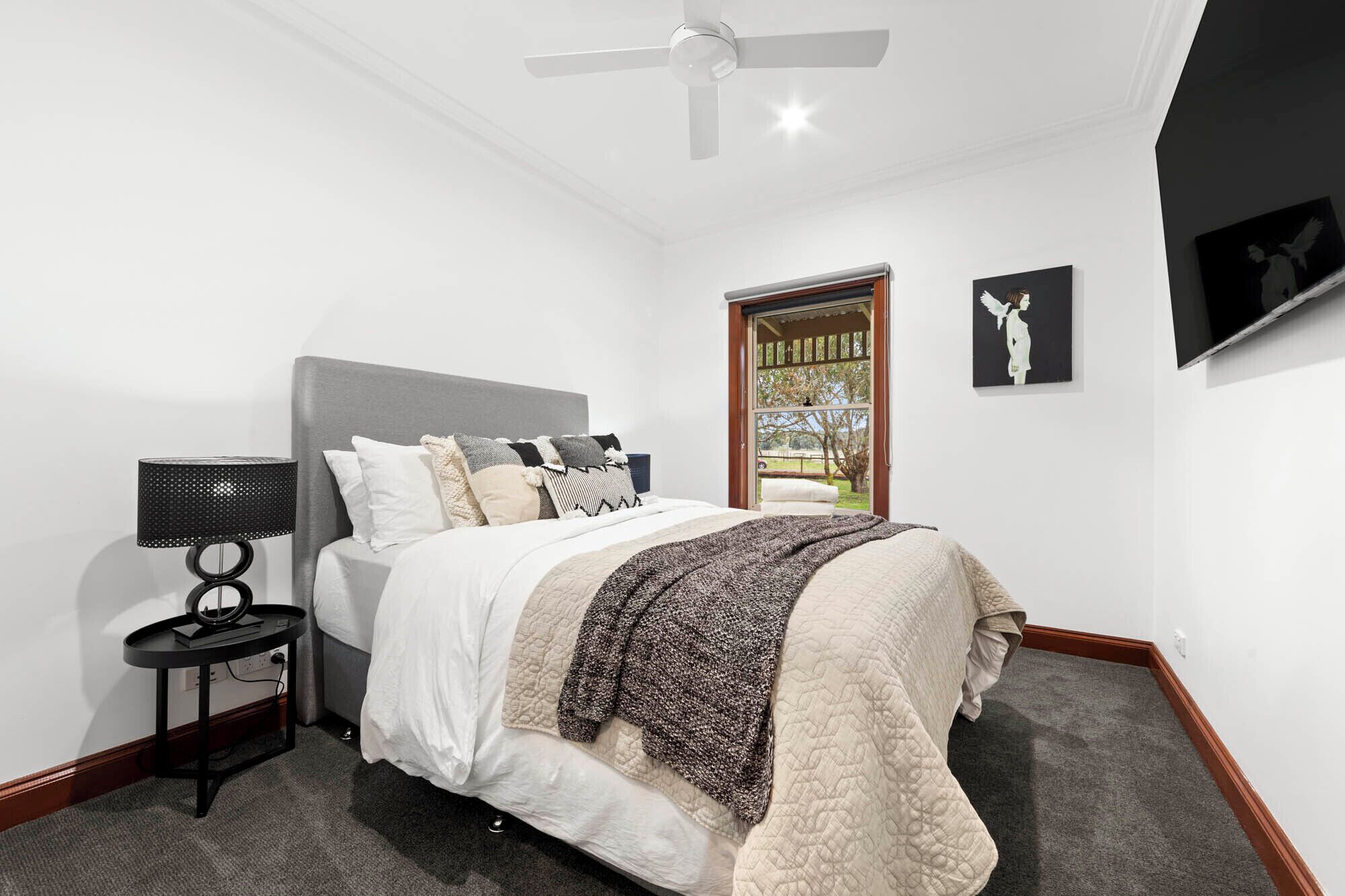 A bedroom with a bed, a nightstand, a ceiling fan, a window with a view, and artwork on the wall.