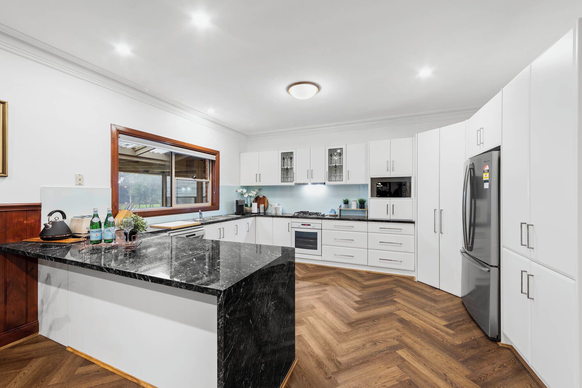 A modern kitchen with white cabinets, a black marble countertop, and a herringbone wood floor.