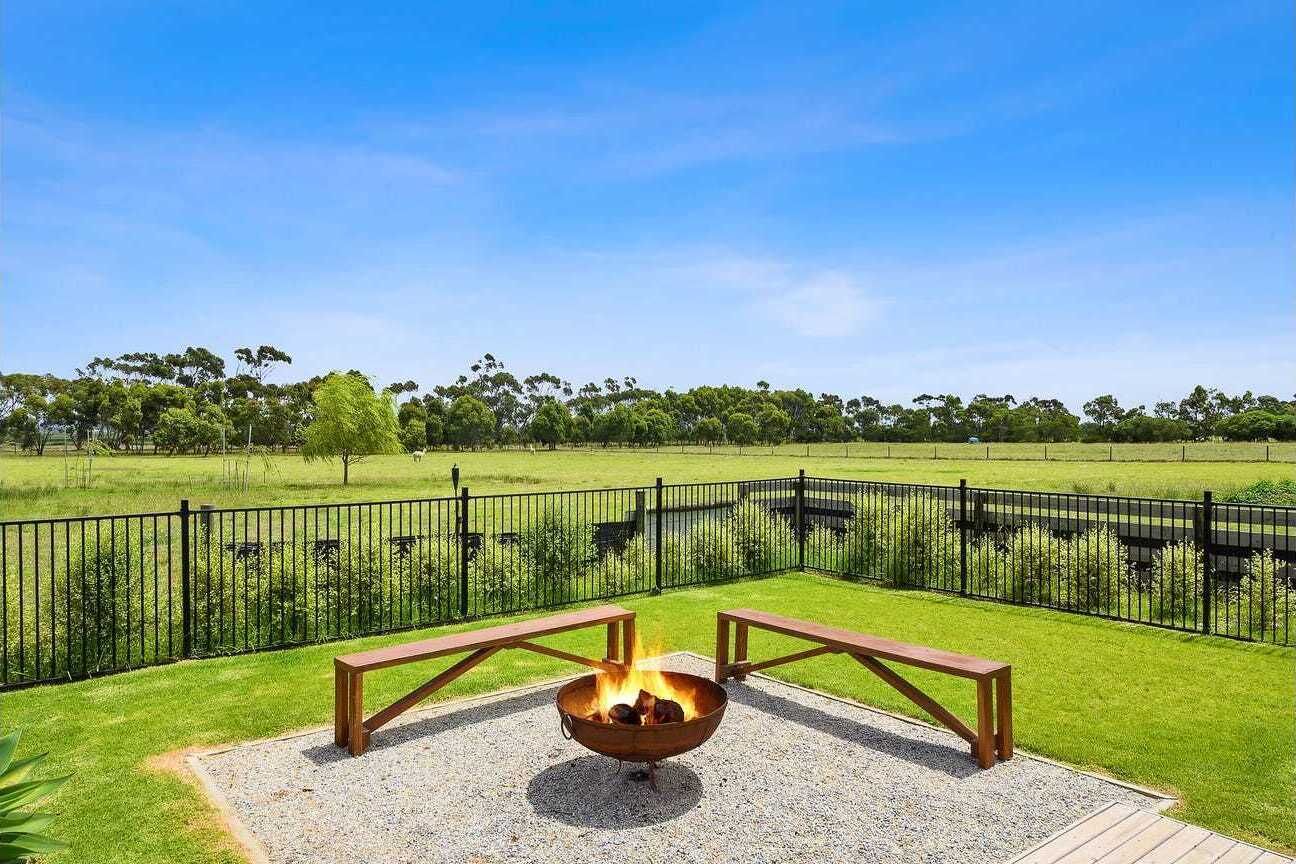 An outdoor seating area with a fire pit and benches, surrounded by a fence and a grassy field.
