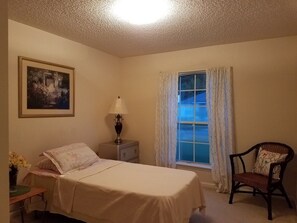 Cozy retreat with a single bed, nightstand, and comfortable chair, plus a window with curtains and a beautiful framed painting.