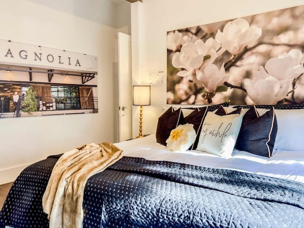 A bedroom with a bed, pillows, a blanket, a wall sign, and a wall art piece. - *Magnolia Masterpiece* | Family Gathering Place (Waco)