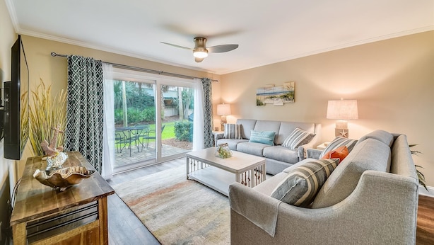 A living room with a sofa, coffee table, and a television. There is a sliding glass door leading to a patio with outdoor furniture. - Sunset Rentals | Island House 125 | Dog Friendly (Hilton Head Island)