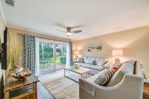 A living room with a sofa, coffee table, and a television. There is a sliding glass door leading to a patio with outdoor furniture.
