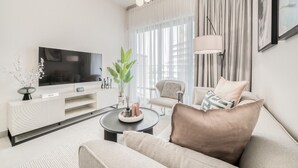 A modern living room with a flat-screen TV, a white entertainment unit, a beige sofa, a coffee table, and a large window with curtains. - Polished 1BR at Binghatti Creek Al Jadaf by Deluxe Holiday Homes (Dubai)