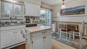 A kitchen with white cabinets, a wooden countertop, a dining table with chairs, a hanging light fixture, and a painting on the wall.
