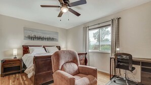 A bedroom with a bed, a wooden nightstand, a desk with a chair, a ceiling fan, and a window with curtains.