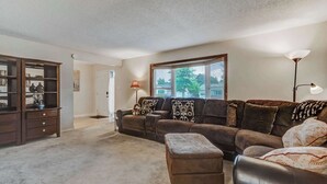 A living room with a sectional sofa, an ottoman, a wooden cabinet, and a large window.