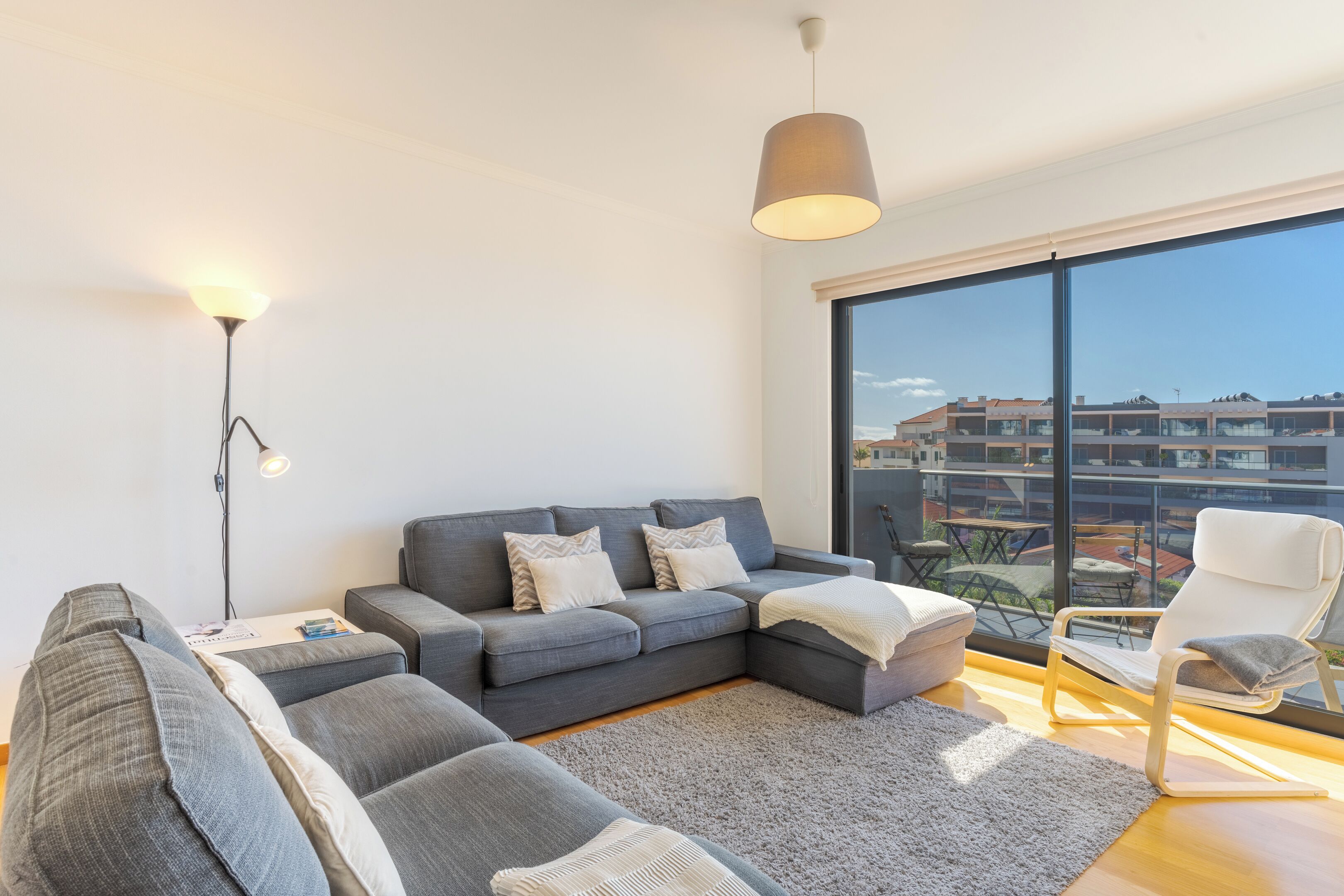 Un soggiorno moderno con un divano modulare grigio, un tavolino con libri, un tappeto grigio e un balcone con vista sugli edifici.