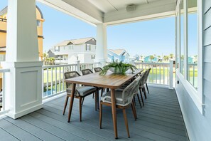 Un patio techado al aire libre con mesa de madera y sillas de mimbre, con vista a una zona residencial con casas y cielo despejado.