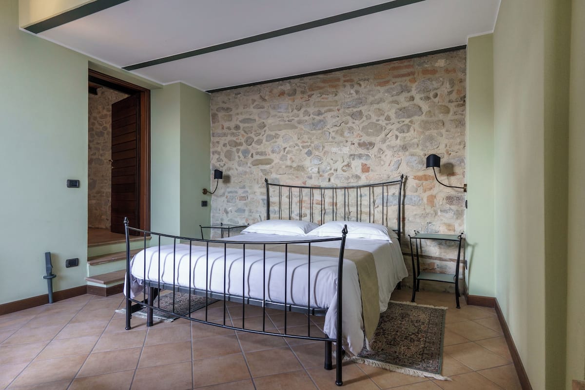 Quarto com parede de pedra, estrutura de cama de metal e mesa de cabeceira com abajur.