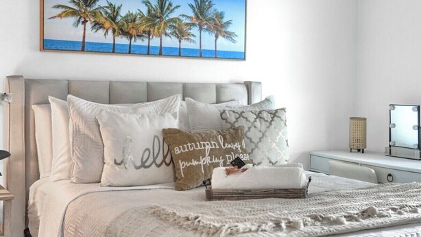 A neatly arranged bedroom with a bed, pillows, a bedside table, a lamp, a mirror, and a painting of palm trees. - Cozy House /MarlinsStadium/ Brickell / Beach / Airport / Port Cruise /Pets (Miami)