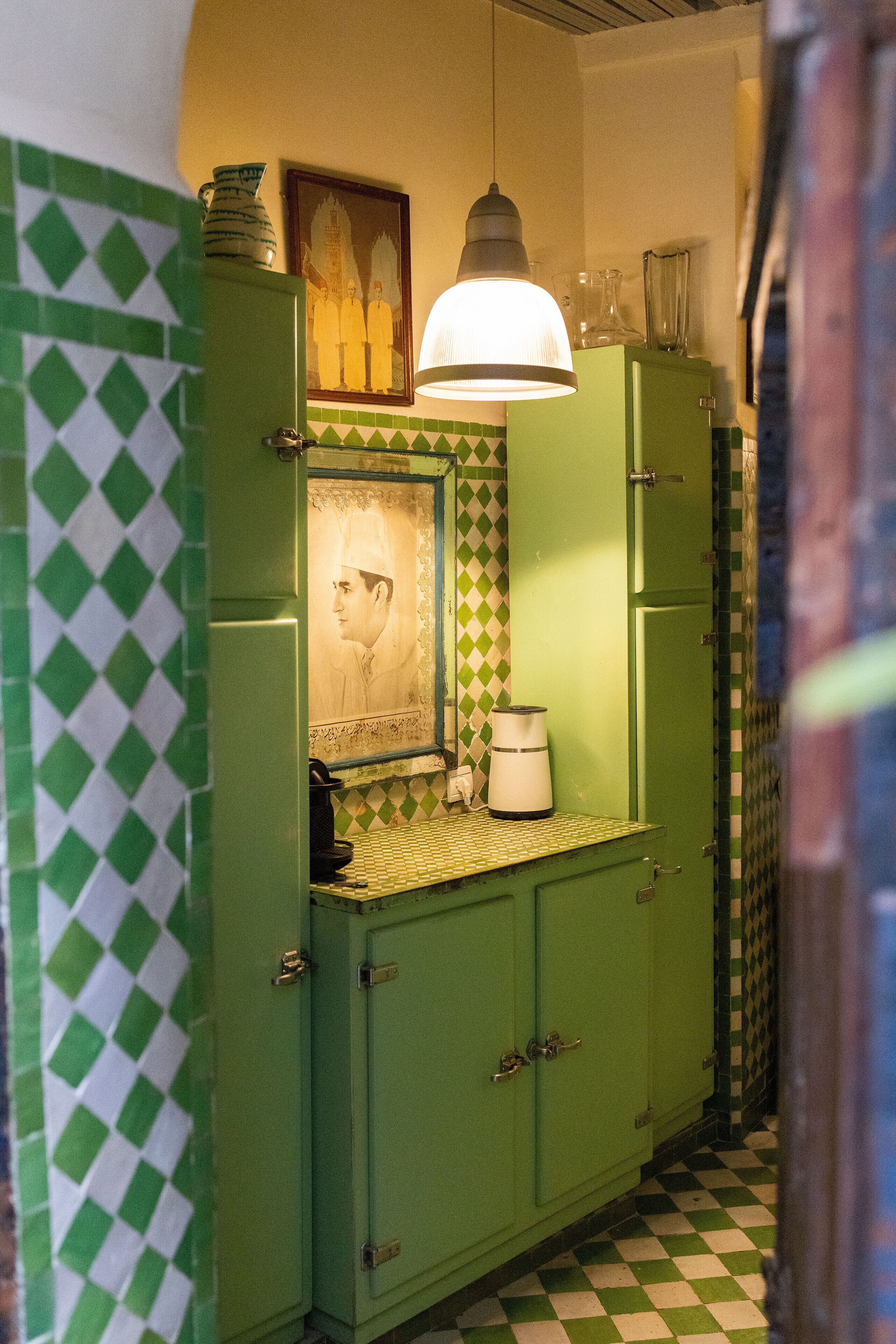 Uma cozinha verde com ladrilhos quadriculados no chão, um lustre pendurado e um quadro na parede.