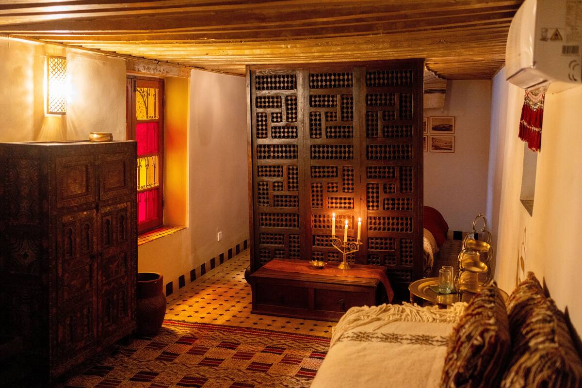 Una habitación tradicional con suelo estampado, un aparador de madera, una cama con cubrecama blanco, un calentador fijado a la pared y un biombo decorativo con una vela.