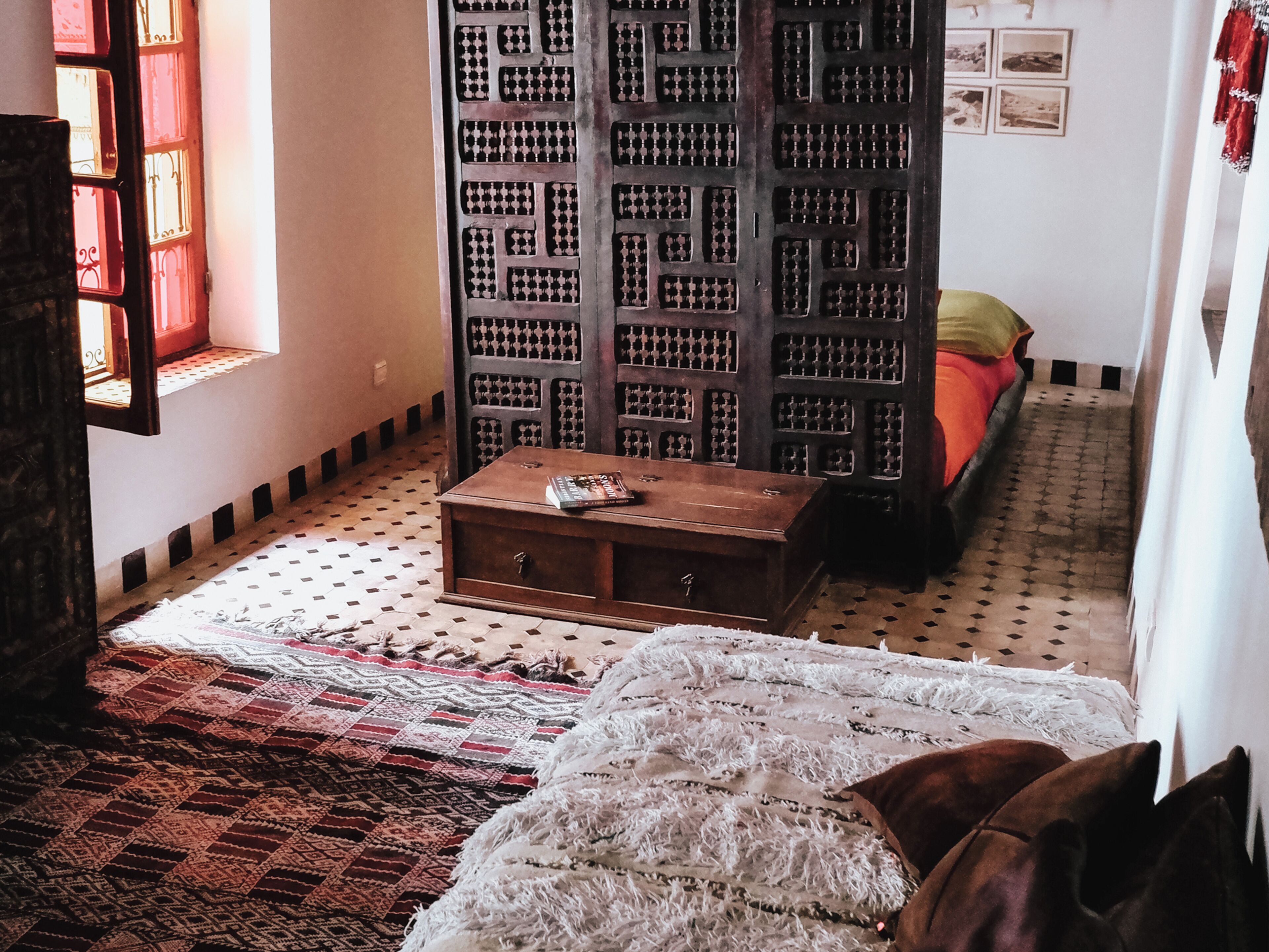 A room with a patterned floor, a wooden chest of drawers, a bed with a red blanket, and a large bookshelf.