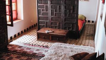 A room with a patterned floor, a wooden chest of drawers, a bed with a red blanket, and a large bookshelf.