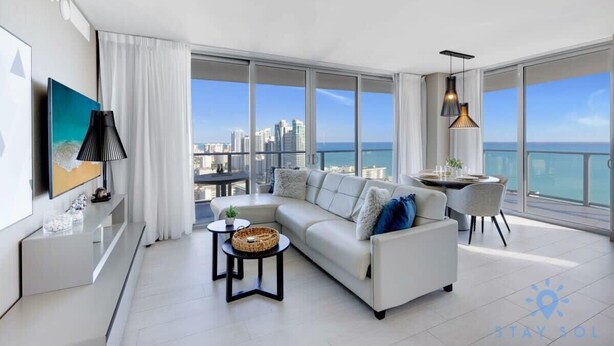 A modern living room with a white sofa, a dining table, and a view of the city and ocean. - Luxury 2Br Front Ocean| Bay View| Amazing Pools (Hollywood)