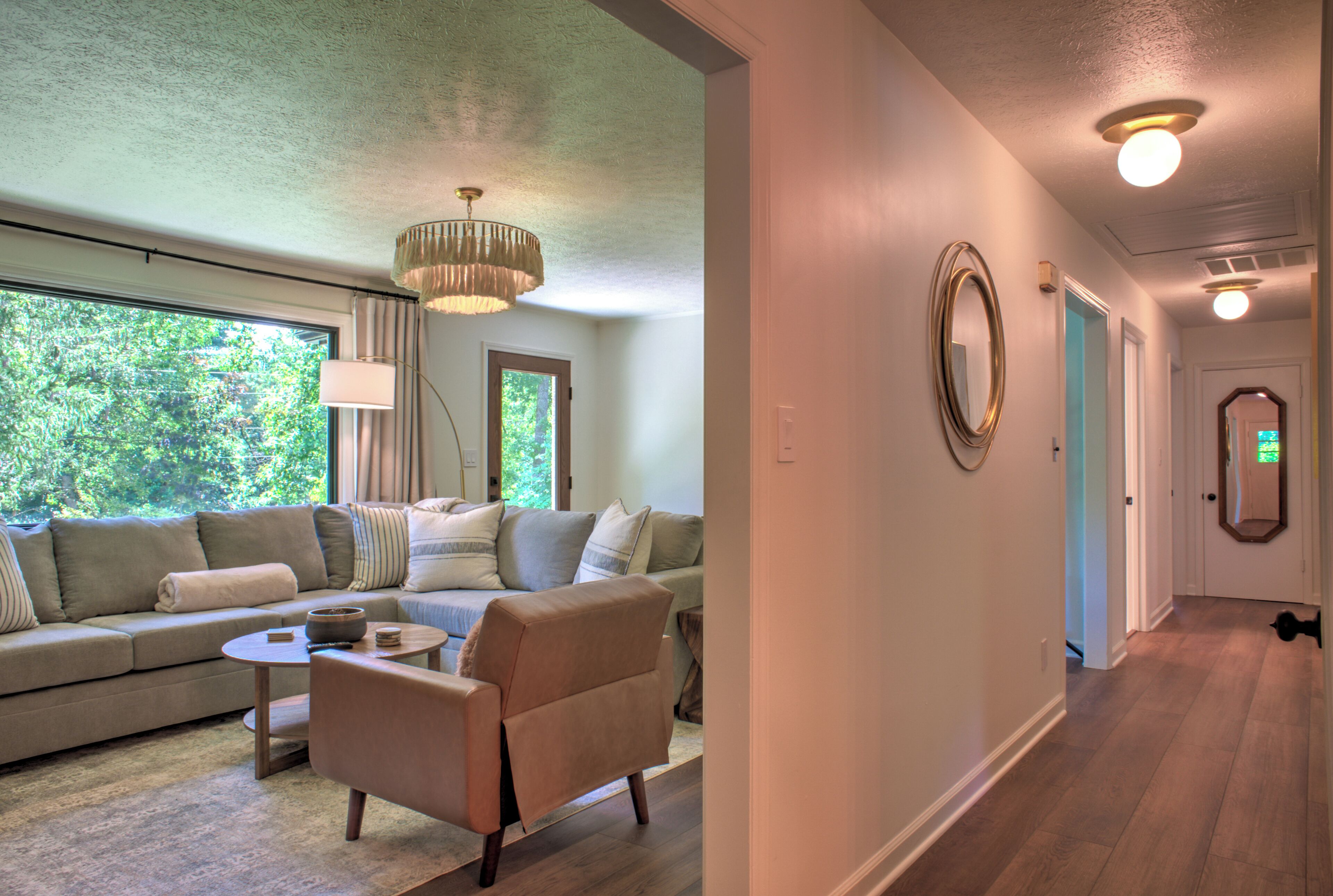 A modern living room with a sectional sofa, a coffee table, and a chair. There is a large window providing natural light and a view of greenery outside.