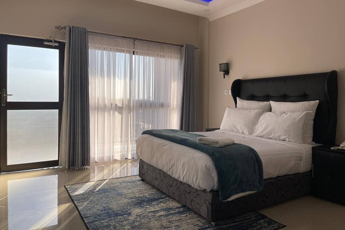 Quarto moderno com uma cama grande, mesa de cabeceira, luminária na parede e vista através de uma porta de vidro.