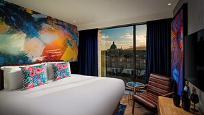 A hotel room with a large bed, a chair, a table, and a view of a cityscape through the window. - NYX Hotel Dublin Portobello (Dublin)