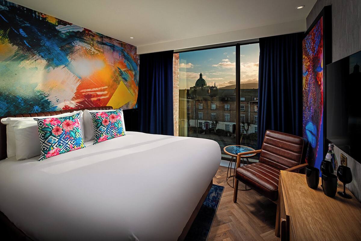 A hotel room with a large bed, a chair, a table, and a view of a cityscape through the window.