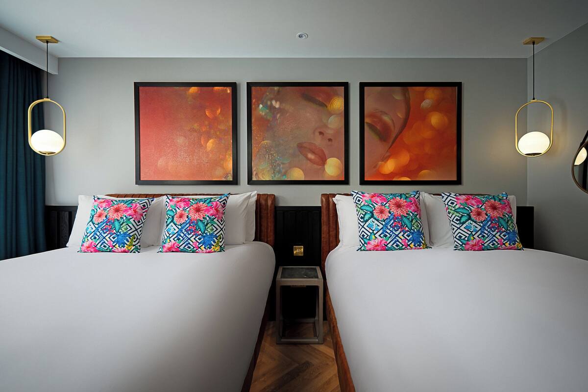 A hotel room with two beds, each with floral-patterned pillows, a small bedside table, and three framed abstract paintings on the wall.