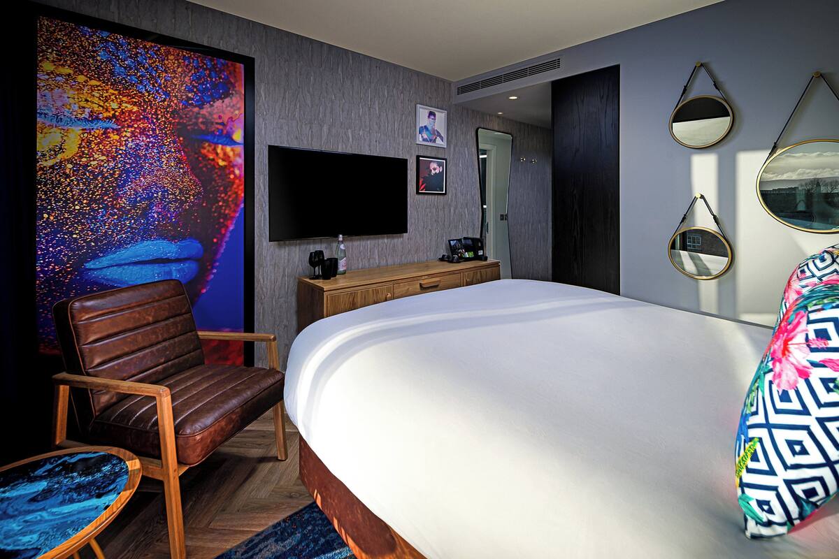 A modern hotel room with a large bed, a wooden nightstand, a flat-screen TV, and a large abstract painting on the wall.