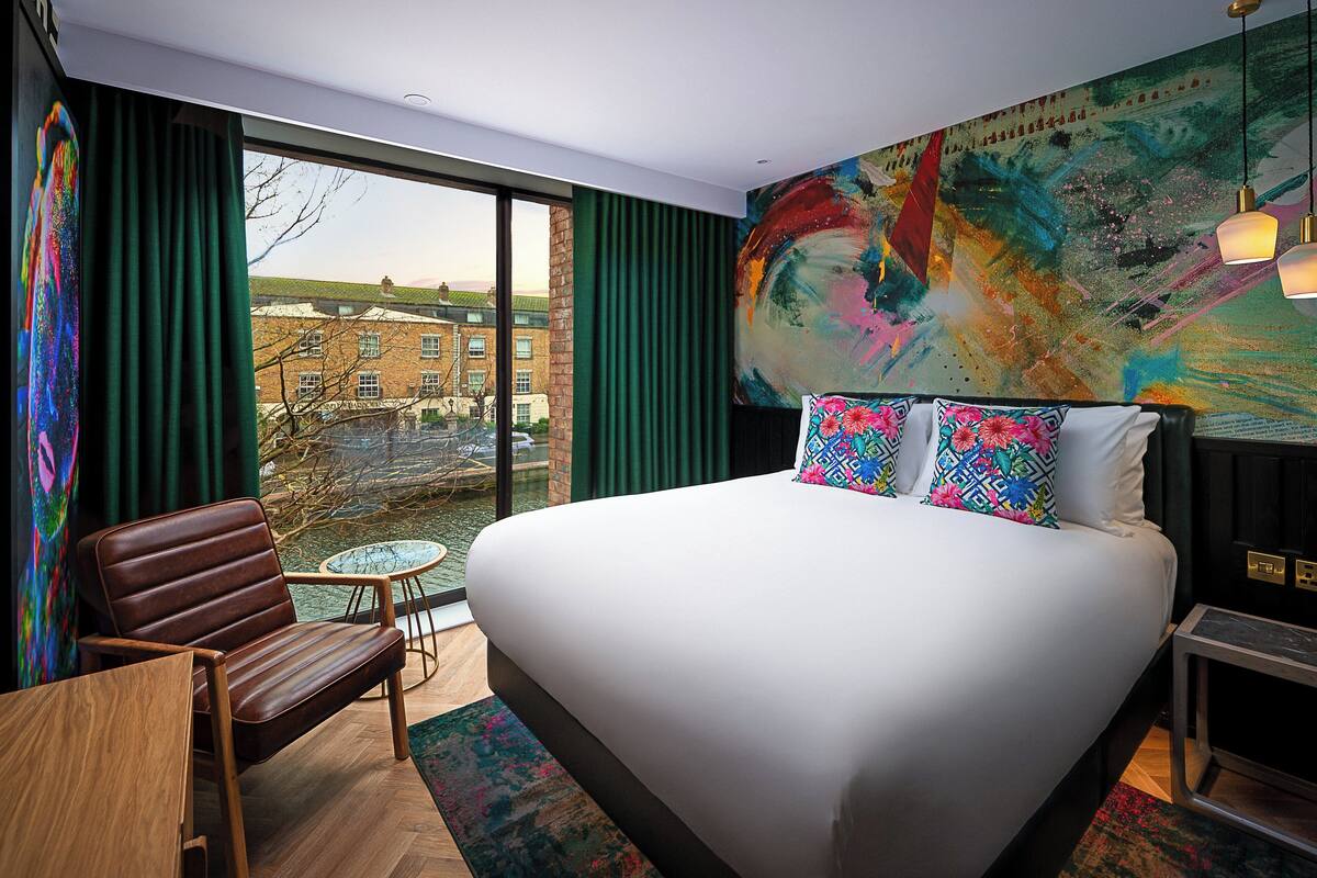 Une chambre d’hôtel moderne avec un grand lit, une vue sur le paysage urbain grâce à de grandes fenêtres du sol au plafond, une petite table ronde et un fauteuil en cuir.