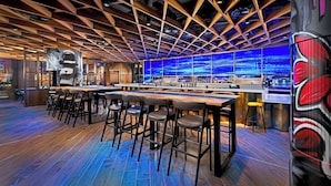 A bar area with wooden ceiling beams, bar stools, and a large screen displaying a blue pattern. - NYX Hotel Dublin Portobello (Dublin)