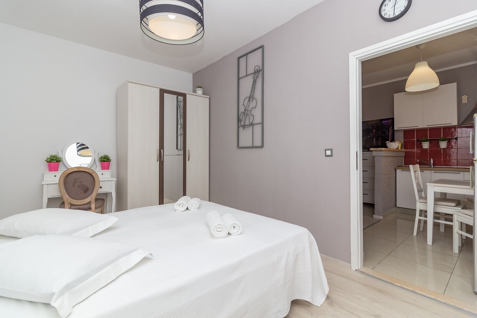 A bedroom with a bed, bedside table, and a mirror. There is a door leading to a bathroom and a kitchenette.