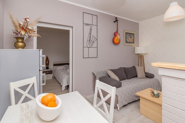Un espace de vie compact comprenant un coin cuisine, un coin repas avec un saladier rempli d’oranges, un canapé gris et une guitare accrochée au mur.