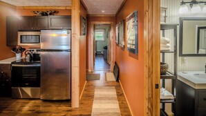 Eine Küche mit Edelstahl-Kühlschrank, Mikrowelle und Herd. Ein Holzboden und ein Flur führen zu einem Badezimmer mit Waschbecken und Spiegel.