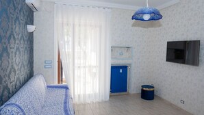 Un salon comprenant un canapé, un téléviseur fixé au mur, un ventilateur de plafond et une fenêtre ornée de rideaux légers.