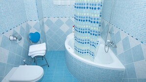 Une salle de bain avec des toilettes blanches, un porte-serviettes bleu en forme de cœur, une baignoire blanche avec un rideau de douche à pois, et des murs et un sol carrelés de bleu.
