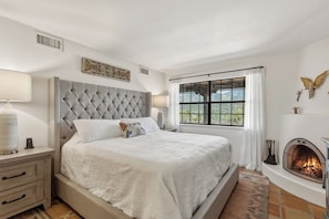 Quarto com uma cama grande, lareira, janela com cortinas e um detalhe decorativo na parede.