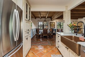Uma cozinha com uma geladeira de aço inoxidável grande, armários de madeira, uma área de refeições com mesa e cadeiras, e vista pelas janelas.