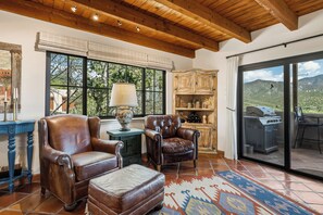 A room with leather chairs, a table, a lamp, a cabinet, and a view of a mountainous landscape through a sliding glass door.