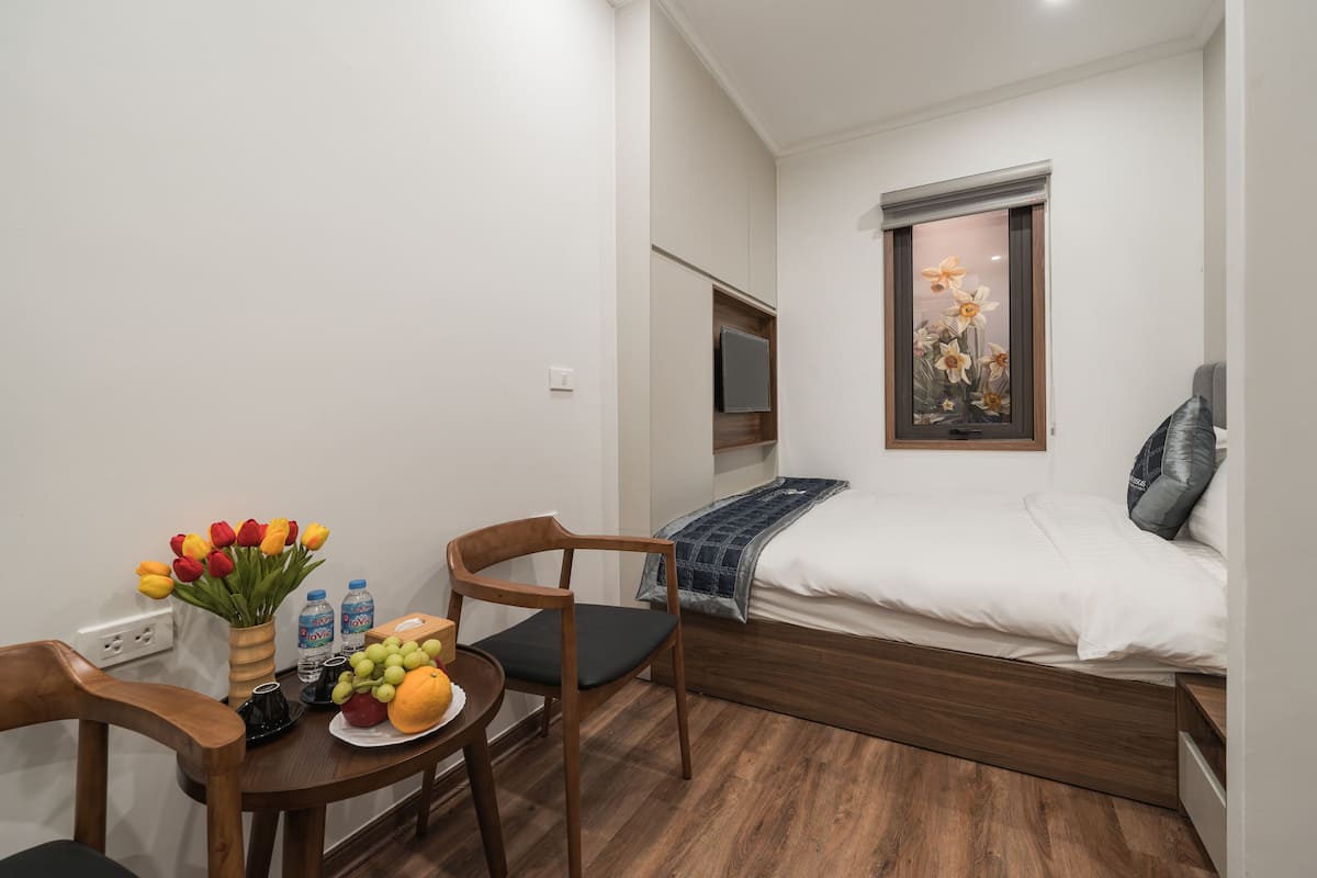 Quarto pequeno e bem arrumado com uma cama, janela com vista, escrivaninha com cadeira e mesa com frutas e flores.