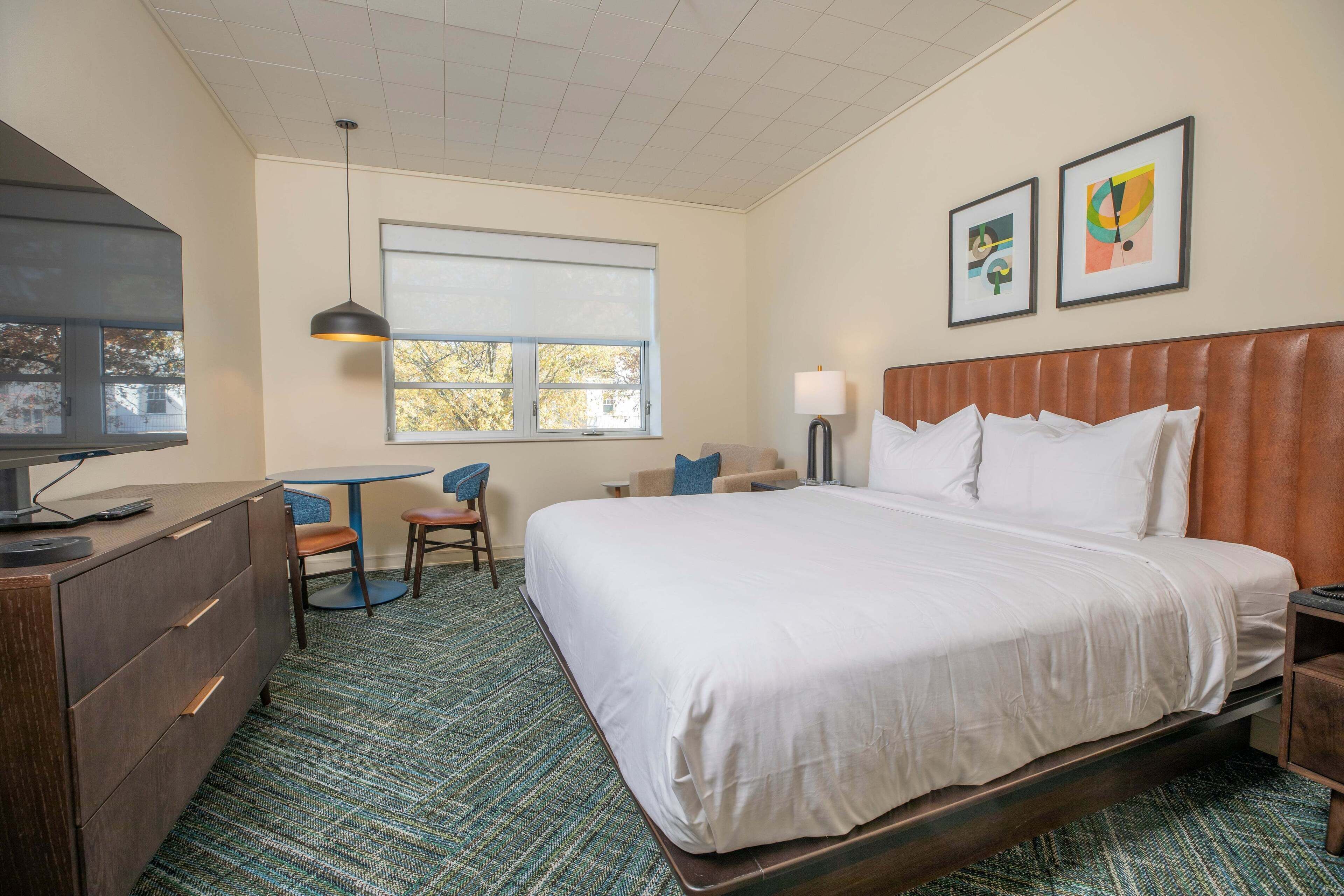 A hotel room with a large bed, a desk with a chair, a television, a window with blinds, and two framed pictures on the wall.