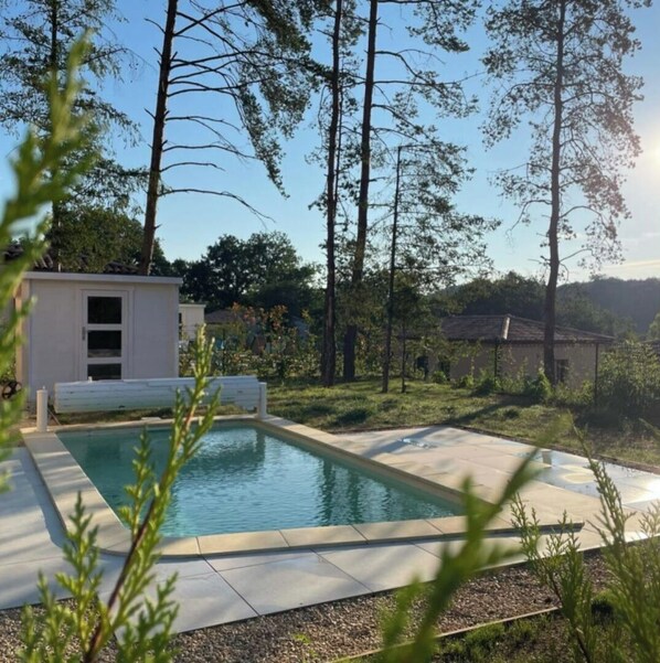 A rectangular pool with a tiled deck, surrounded by a grassy area with trees and a small white building in the background.