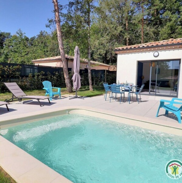 Une piscine entourée d’arbres et d’un bâtiment à toit de tuiles, avec des chaises longues et une table à proximité.