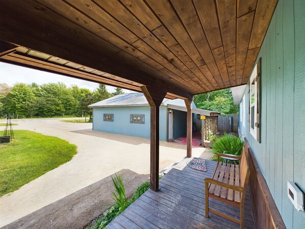 A covered porch with a wooden bench, a small table, and a blue house with a metal roof in the background.