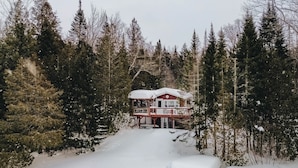 Eine Holzhütte mit überdachter Terrasse und einem verschneiten Weg, der davor führt, umgeben von dichten Nadelbäumen.