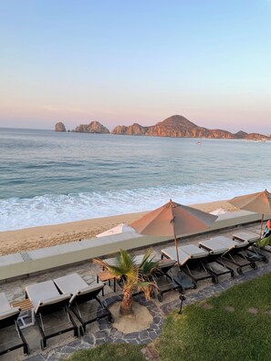Eine Strandlounge mit Liegestühlen und Sonnenschirmen, einer Palme und Blick auf das Meer und vorgelagerte Inseln.