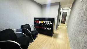 A hallway with a black sign reading "ONHOSTEL GUARUTHOS" and "SANTA SE EM CASA". There are black chairs lined up against the wall. - On Hostel gru unidade 2 traslado café  (Guarulhos)