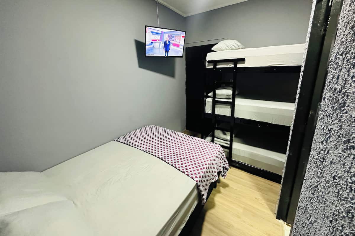 Ett litet rum med en enkel säng, och i bakgrunden syns en våningssäng. En tv är monterad på väggen och golvet är täckt av ett grått heltäckningsmatta.